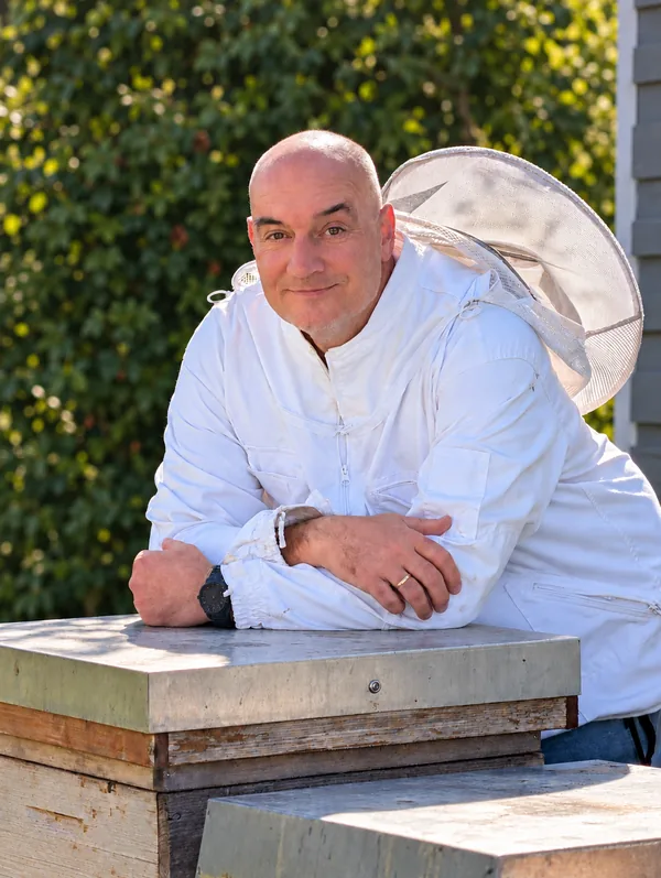 Michael Benoist im weißen Imkeranzug mit zurückgeschobenem Schleierhut, auf eine Bienenbeute gelehnt